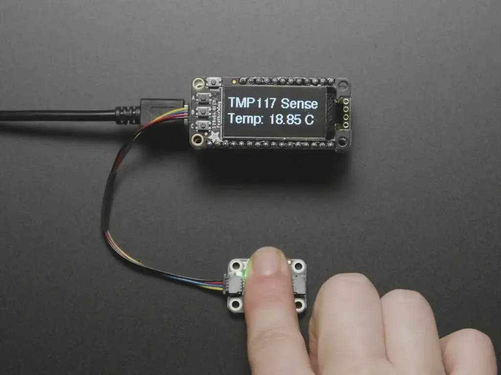 A finger warms a TMP117 breakout connected to a controller and temperature display